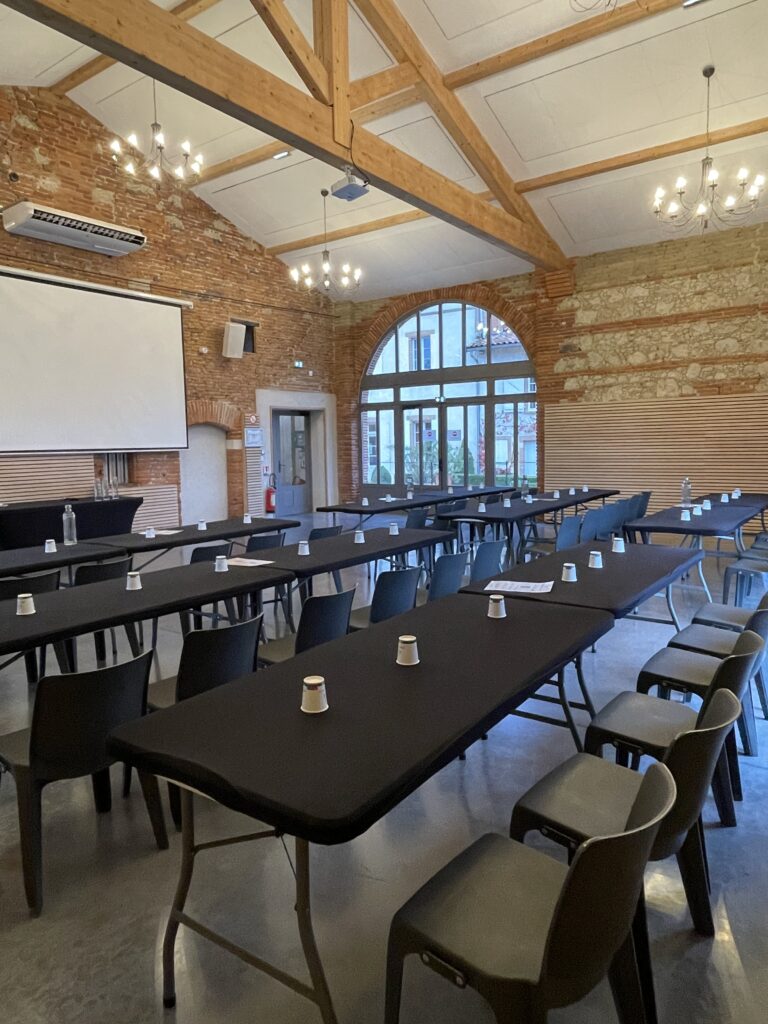 Salle de réunion configurée en U avec tables noires, chaises et écran de projection, dans un cadre chaleureux en briques.